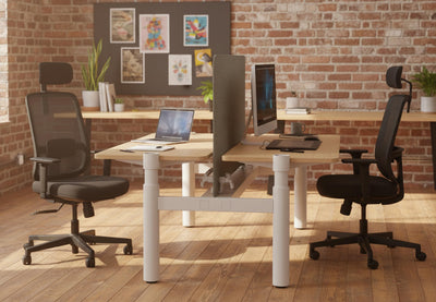Fokus Ergonomic High-Back Chairs in black shown in a modern office interior, featuring two chairs positioned at work desks within a space with exposed brick walls, wooden flooring, natural light and greenery, highlighting contemporary ergonomic office seating and profesional workplace design.