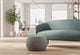 Edgar Plush Curve Pouf positioned at the centre of a modern office breakout room, styled in front of a Keebu Breakout 2 Seater Sofa. Set within a bright workplace interior featuring white walls, large windows, natural light, greenery and wooden flooring with office desks and chairs visible in the background, highlighting contemporary breakout and collaborative seating design.