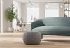 Edgar Plush Curve Pouf positioned at the centre of a modern office breakout room, styled in front of a Keebu Breakout 2 Seater Sofa. Set within a bright workplace interior featuring white walls, large windows, natural light, greenery and wooden flooring with office desks and chairs visible in the background, highlighting contemporary breakout and collaborative seating design.