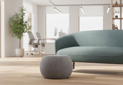 Edgar Plush Curve Pouf positioned at the centre of a modern office breakout room, styled in front of a Keebu Breakout 2 Seater Sofa. Set within a bright workplace interior featuring white walls, large windows, natural light, greenery and wooden flooring with office desks and chairs visible in the background, highlighting contemporary breakout and collaborative seating design.