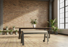 Modern industrial office with a dark wood table and light beige bench, both central to the foreground. A white vase with fresh flowers sits on the table, while exposed brick, concrete pillars, wooden floors, and potted plants fill the background, illuminated by large windows.