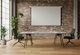 Modern industrial meeting room with a large conference table, two black office chairs, a projector screen on an exposed brick wall, and abundant natural light from large windows. Features hardwood floors and various green plants, suitable for collaborative workspaces